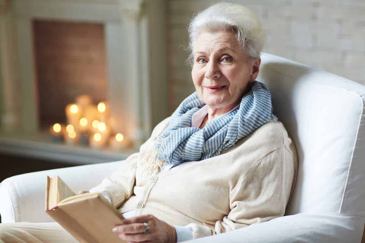 Smiling retired woman seated on a white chair reading a book with lit candles in the background.