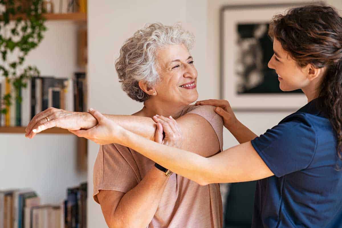Older woman smiling as a younger woman helps her stretch her shoulder.