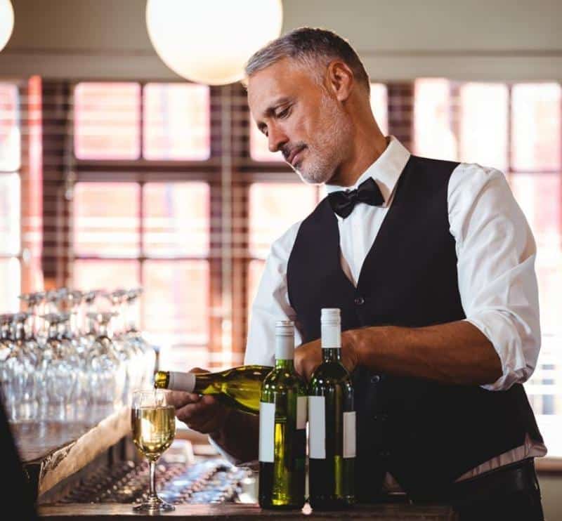 Handsome waiter in black and white attire pouring glasses of white wine.