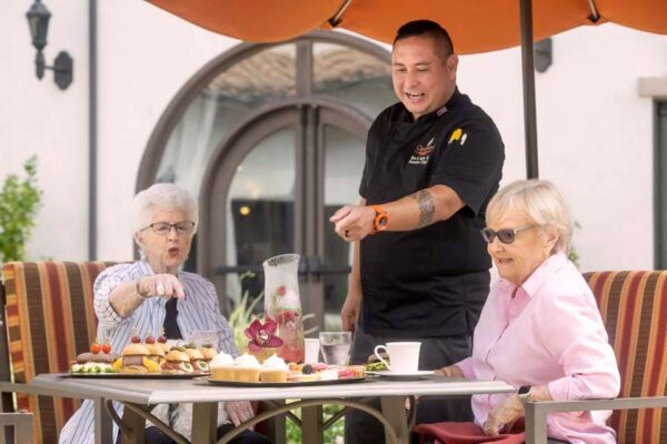 Two Crestavilla residents enjoying an al fresco lunch on the patio with Chef Bert explaining some of the menu items.